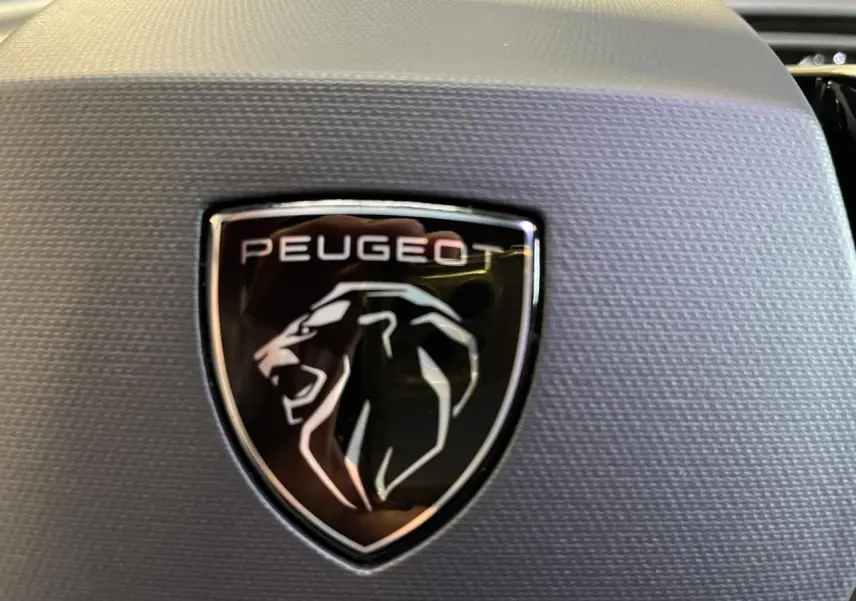 Gros plan sur le logo Peugeot noir et argenté sur fond texturé gris, intérieur du volant du 3008 GT hybride blanc.
