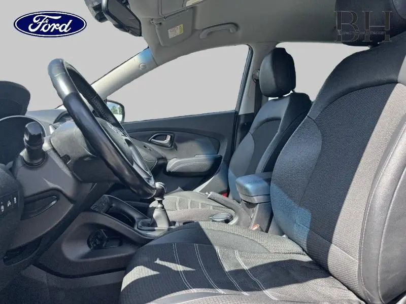 Intérieur avant côté gauche d’un Hyundai ix35 2014, sièges tissu noirs avec accoudoir central et volant cuir.