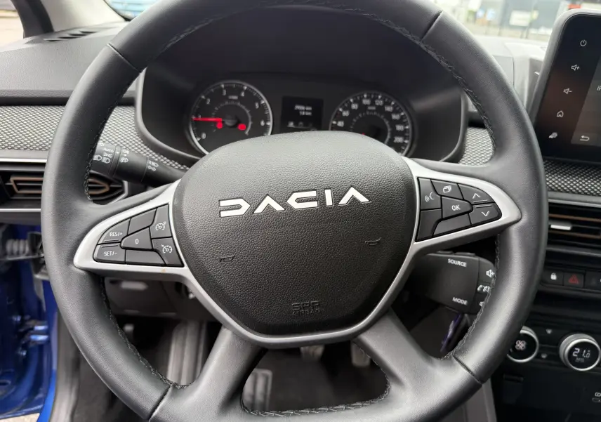 Vue rapprochée du volant noir de la Dacia Sandero Stepway 2025 avec commandes intégrées et tableau de bord en arrière-plan.
