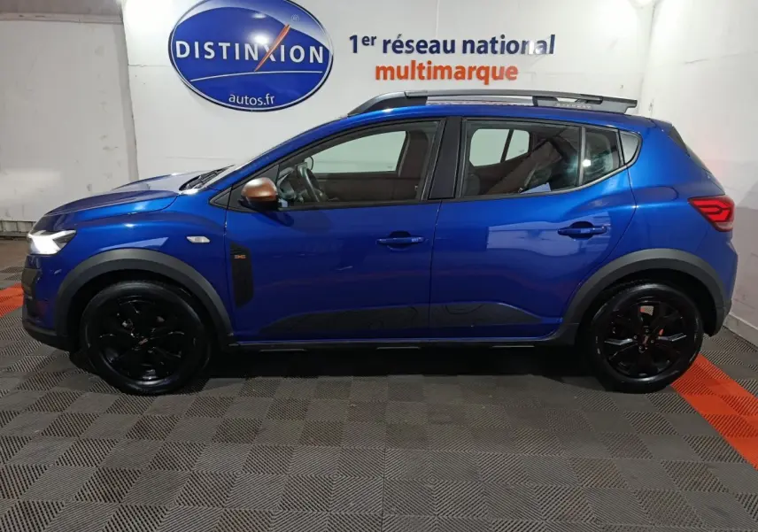 Profil côté gauche d'une Dacia Sandero Extreme+ TCe 110 bleu 2024 avec jantes noires et barres de toit assorties.