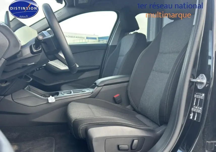 Vue intérieure côté conducteur de la BMW Série 1 120da 2024 noire, sièges tissu anthracite avec accoudoir central.