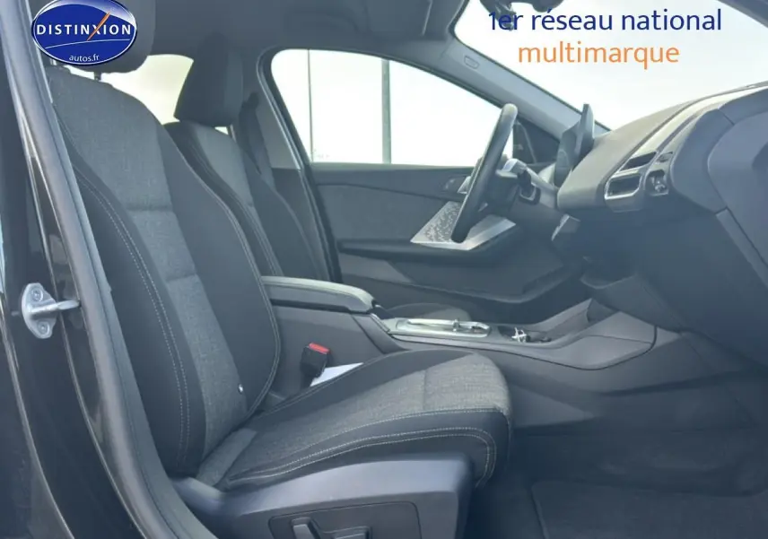 Intérieur avant droit de la BMW Série 1 noire 2024, sièges tissu anthracite avec accoudoir central et tableau de bord moderne.