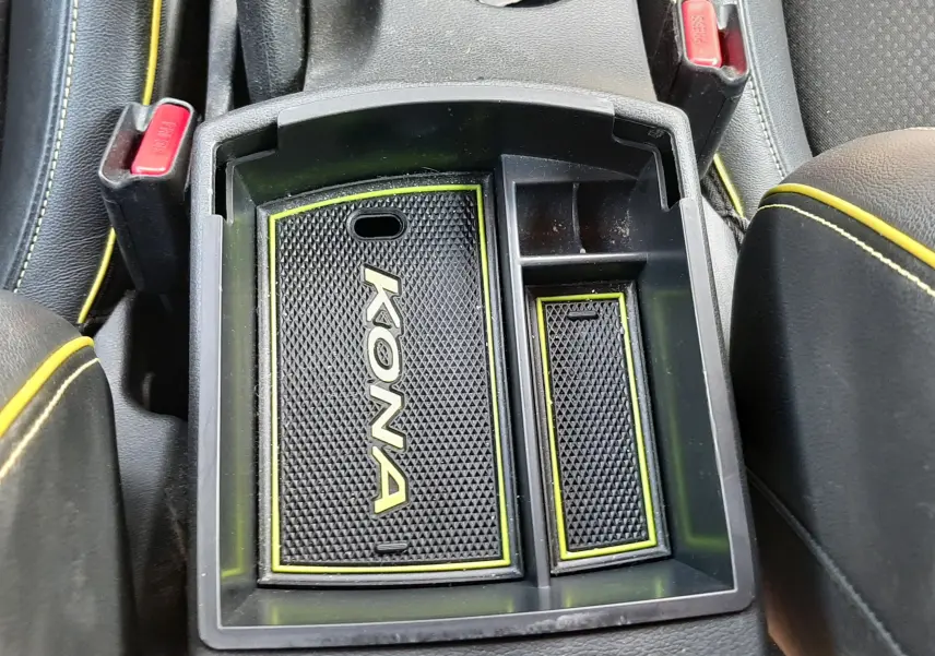 Vue plongeante sur le rangement central intérieur du Hyundai Kona gris avec tapis antidérapant marqué "KONA" et surpiqûres jaunes.