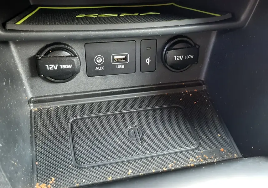 Gros plan sur la console centrale du Hyundai Kona gris, montrant prises 12V, USB, AUX et chargeur sans fil Qi avec traces de saleté.