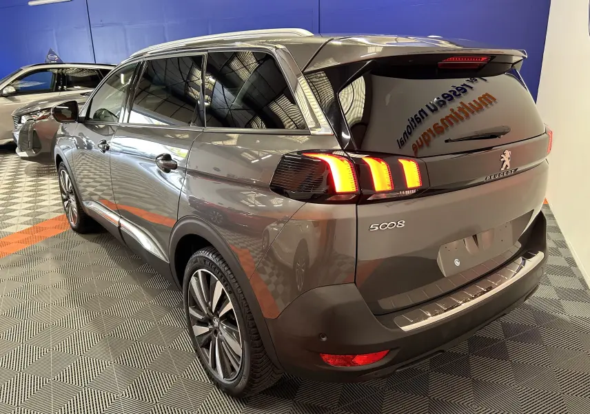 Peugeot 5008 gris vue 3/4 arrière droit dans un showroom, feux arrière allumés et jantes alliage visibles.