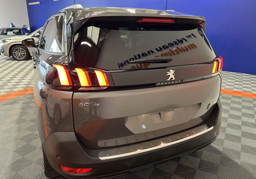 Vue 3/4 arrière d'un Peugeot 5008 gris avec feux arrière allumés dans un showroom intérieur.