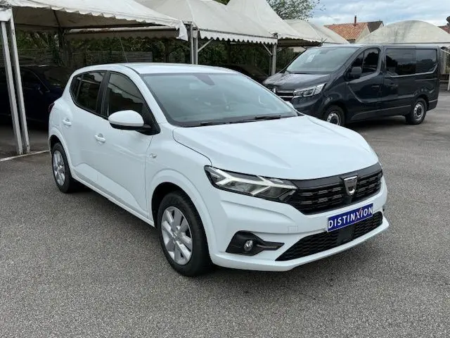Dacia Sandero blanc glacier vue 3/4 avant droit, calandre noire et feux LED, stationnée sur parking extérieur.