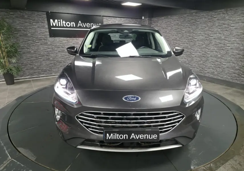 Vue avant d'un Ford Kuga gris métallisé 2020 avec calandre chromée et phares allumés dans un showroom.