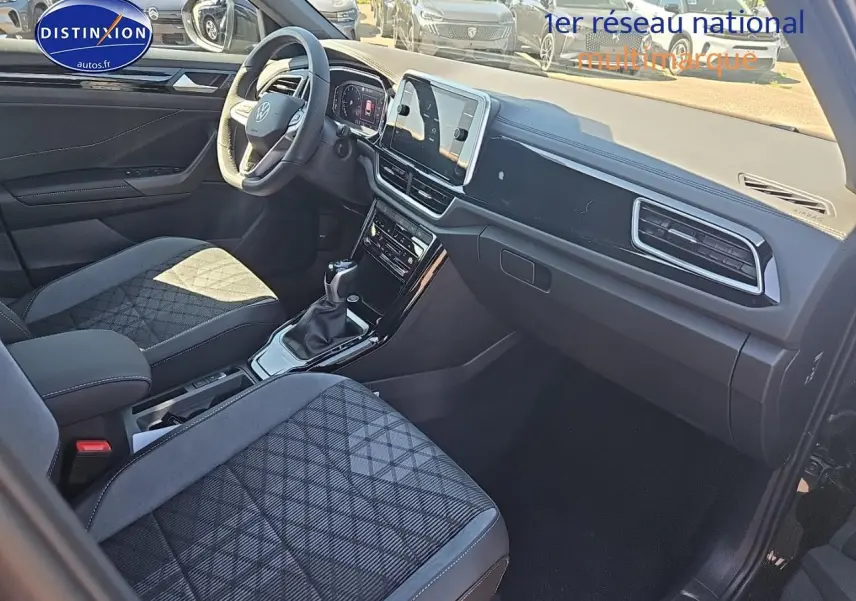 Intérieur du Volkswagen T-Roc 2025 vu côté passager, sièges tissu noir à motifs et tableau de bord moderne.