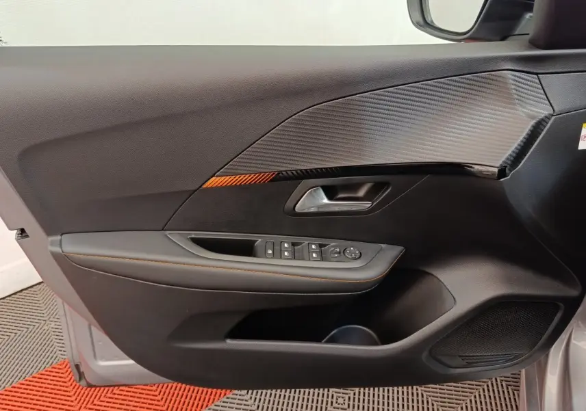 Vue intérieure côté gauche de la porte avant gris clair d’une Peugeot 208 Electrique ROADTRIP avec insert décor carbone et surpiqûres orange.