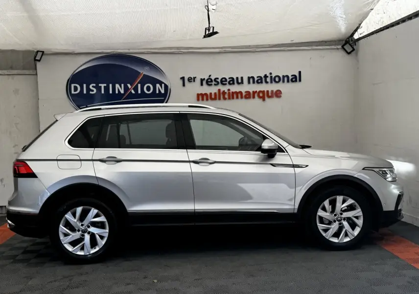 Vue latérale droite d'un Volkswagen Tiguan gris clair 2021 dans un showroom avec logo Distinxion en arrière-plan.