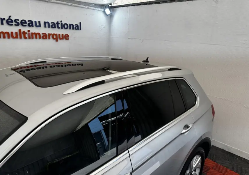 Vue 3/4 arrière droite d’un Volkswagen Tiguan gris clair avec toit ouvrant et barres de toit chromées.