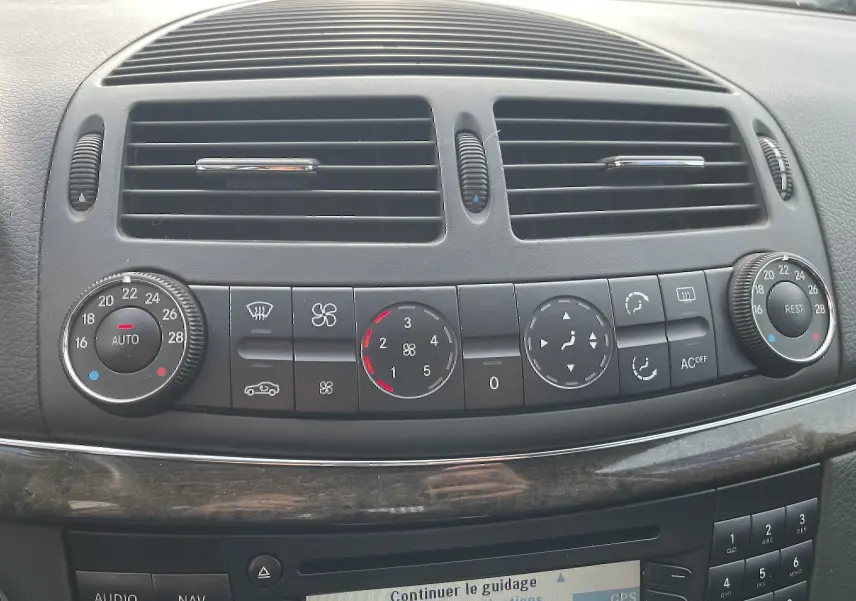 Gros plan sur la console centrale noire de la Mercedes Classe E 320 CDI 2003, montrant les commandes de climatisation et les aérateurs.