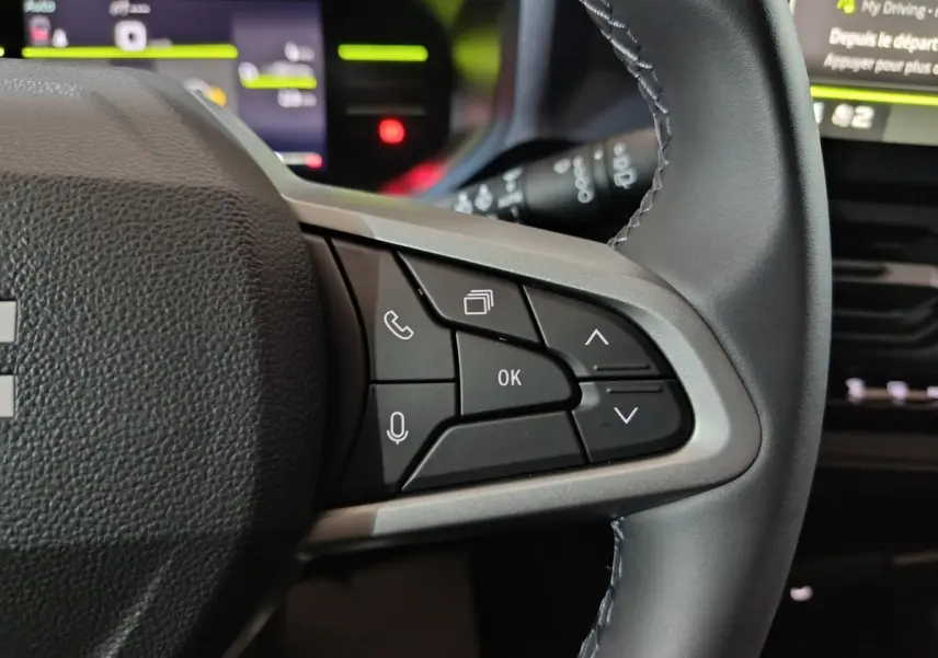 Gros plan sur les commandes multifonctions à droite du volant gris foncé du Dacia Bigster, avec affichage numérique flou en arrière-plan.