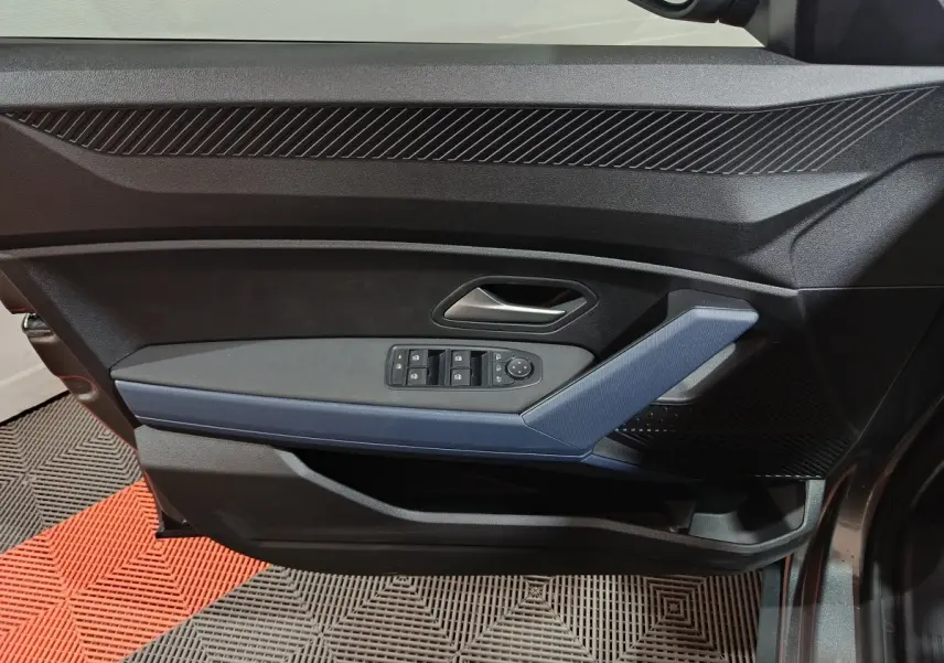 Vue intérieure côté gauche de la porte avant grise foncée du Dacia Bigster avec commandes électriques des vitres et poignée bleue.