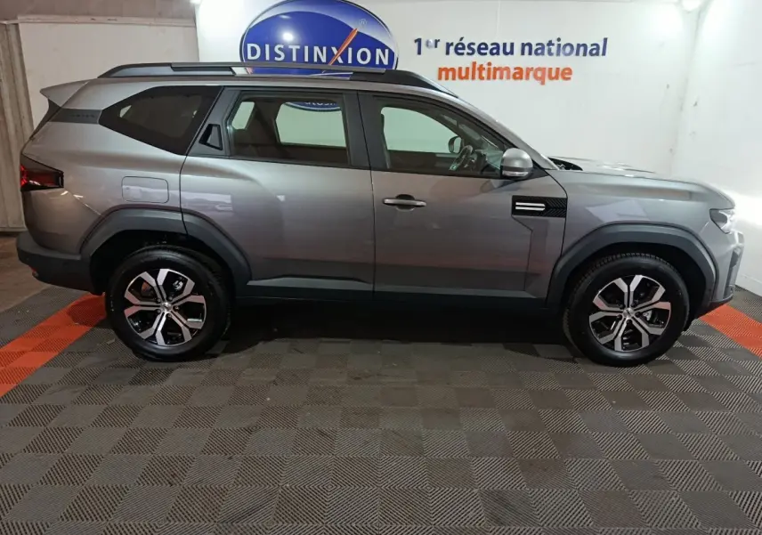Vue de profil côté droit du SUV Dacia Bigster gris foncé avec jantes alliage et barres de toit noires.