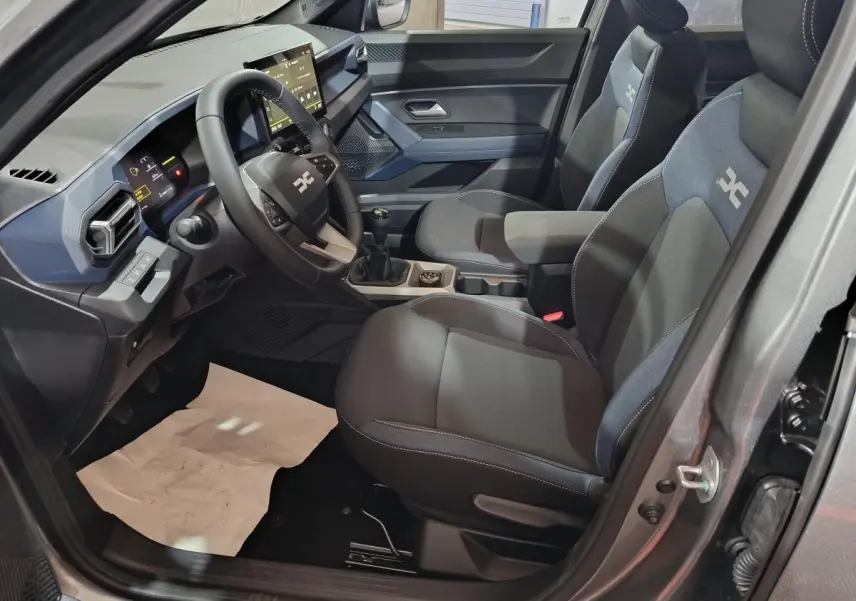 Vue intérieure côté conducteur du Dacia Bigster gris foncé, montrant le tableau de bord moderne et les sièges tissu avec logo.