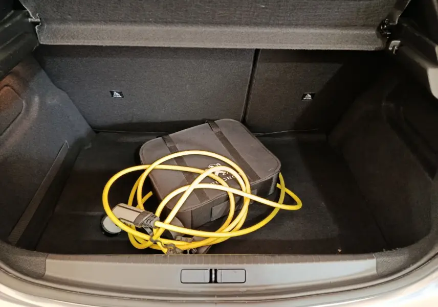 Coffre ouvert d’une Peugeot 208 électrique gris clair avec câble de recharge jaune enroulé dans un sac noir.