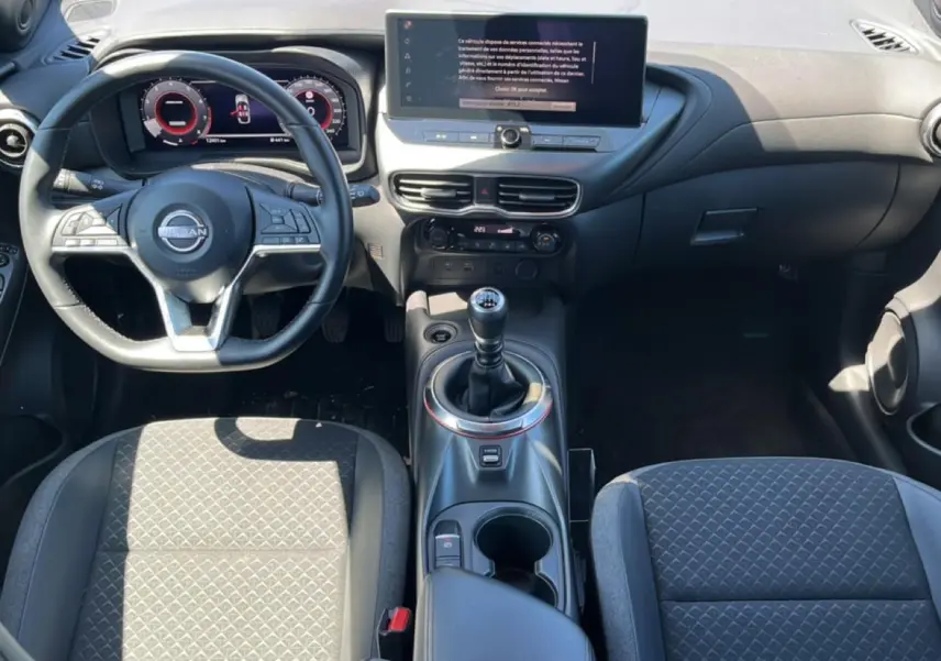 Vue intérieure avant du Nissan Juke 2025 gris, volant cuir multifonction et levier de vitesse manuel au centre.