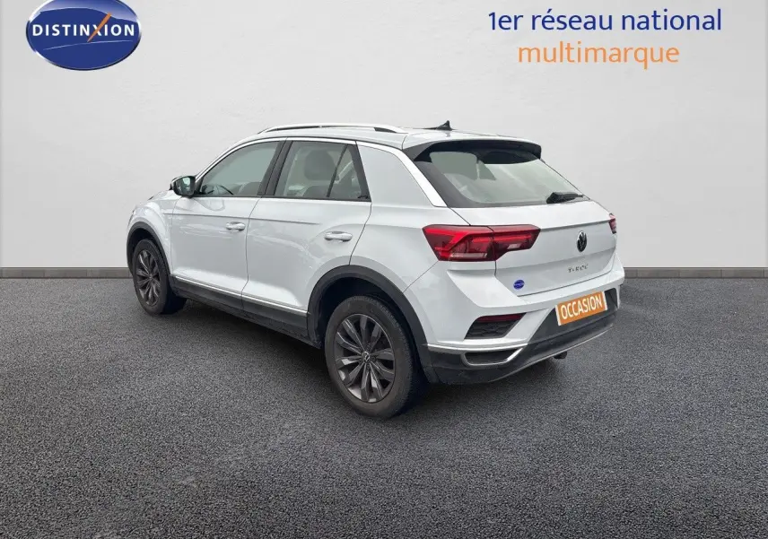 Volkswagen T-Roc blanc argenté vu en 3/4 arrière droit, avec jantes foncées et feux arrière allumés.