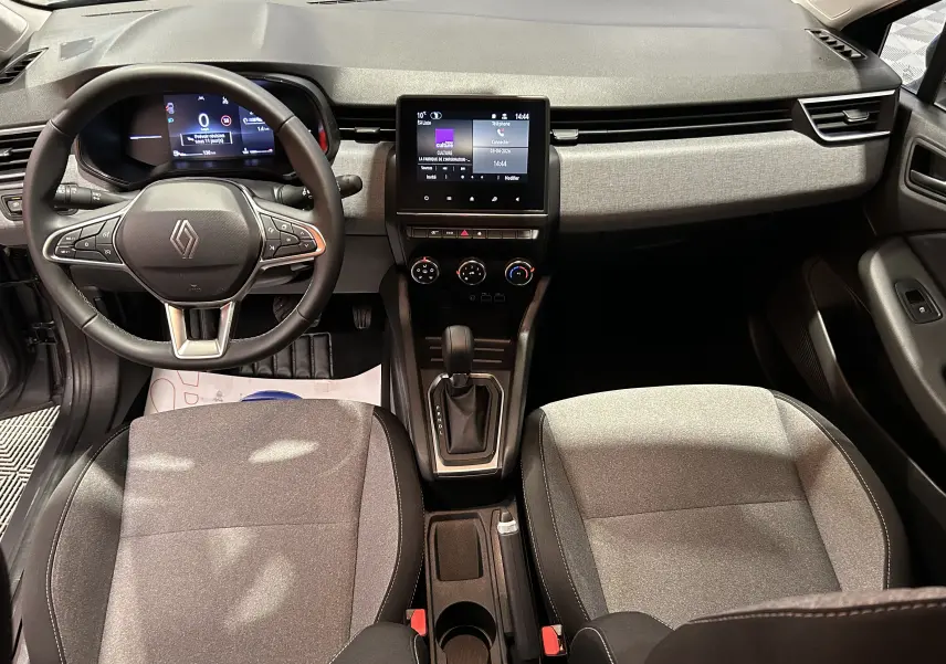 Vue intérieure avant de la Renault Clio V 2025, tableau de bord gris clair avec écran tactile central et volant cuir noir.