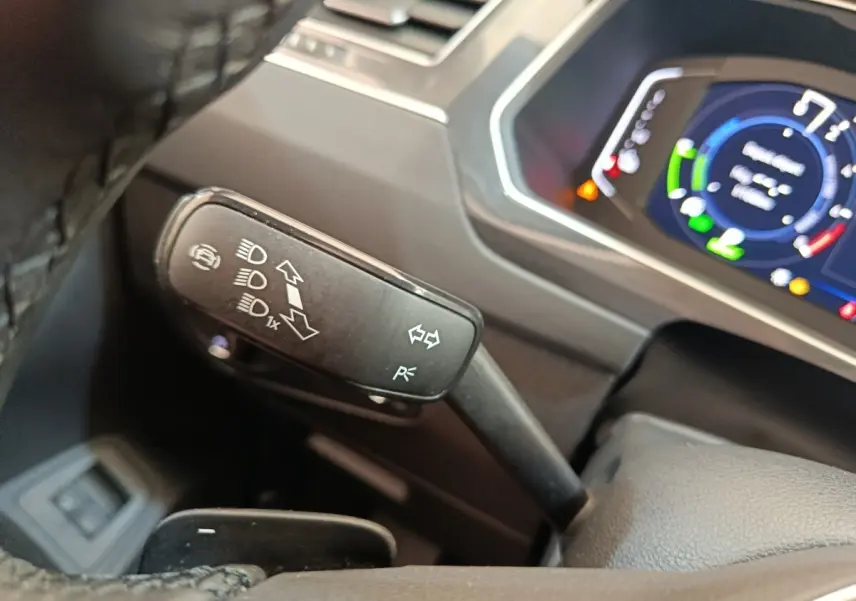 Gros plan sur la manette de clignotant noire à gauche du volant, avec tableau de bord numérique coloré en arrière-plan.