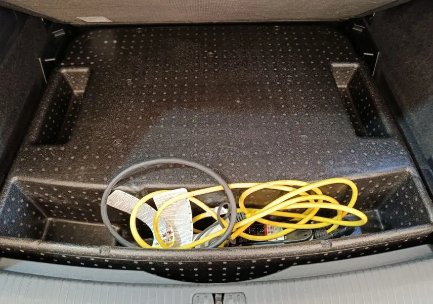 Vue plongeante sur le rangement sous plancher de coffre du Volkswagen Tiguan noir, avec câble de recharge jaune et noir visible.