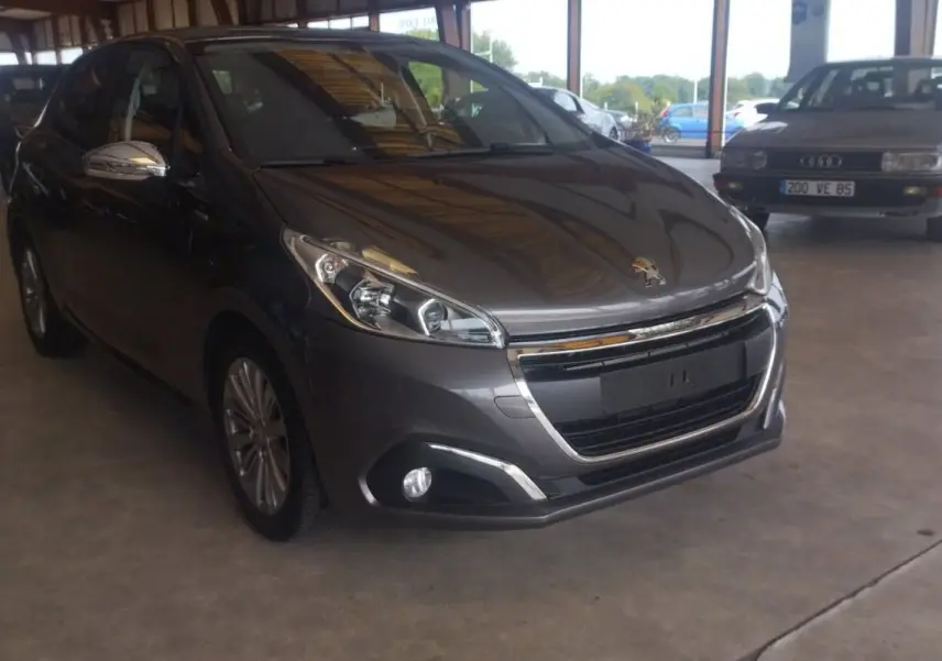 Peugeot 208 gris métal vue 3/4 avant droit, avec calandre chromée et phares modernes, en intérieur d'exposition.