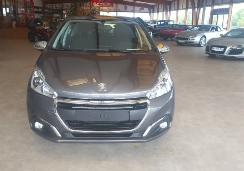 Vue avant d'une Peugeot 208 gris métal 2023 exposée en intérieur avec calandre chromée et phares allumés.