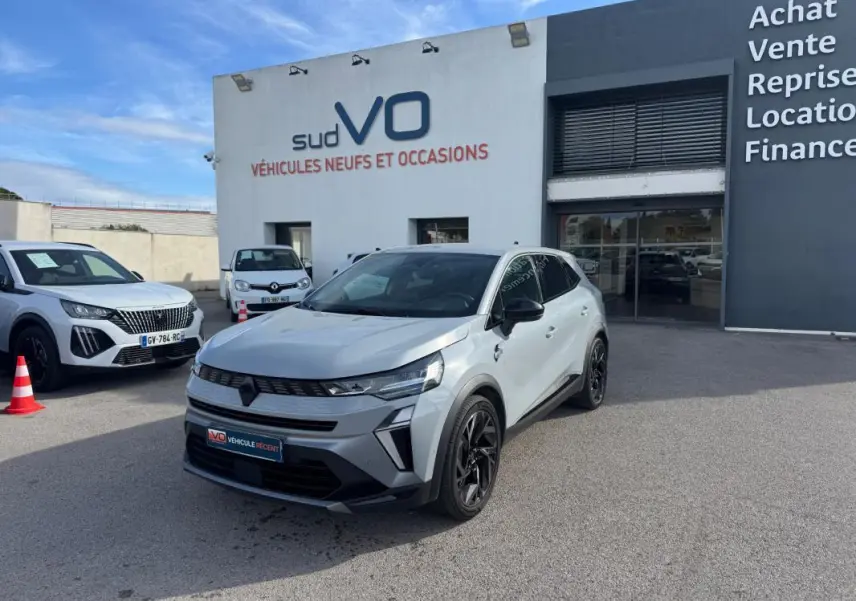 Renault Symbioz gris clair en 3/4 avant droit, jantes alliage noires et toit noir, devant un concessionnaire.