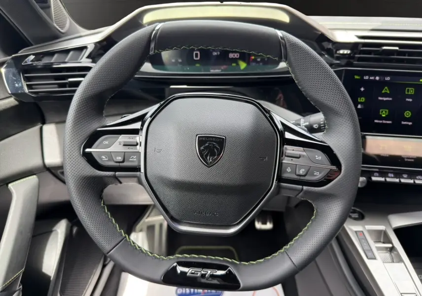 Vue rapprochée du volant cuir noir avec surpiqûres vertes et logo GT du Peugeot 308 2026, tableau de bord digital visible.