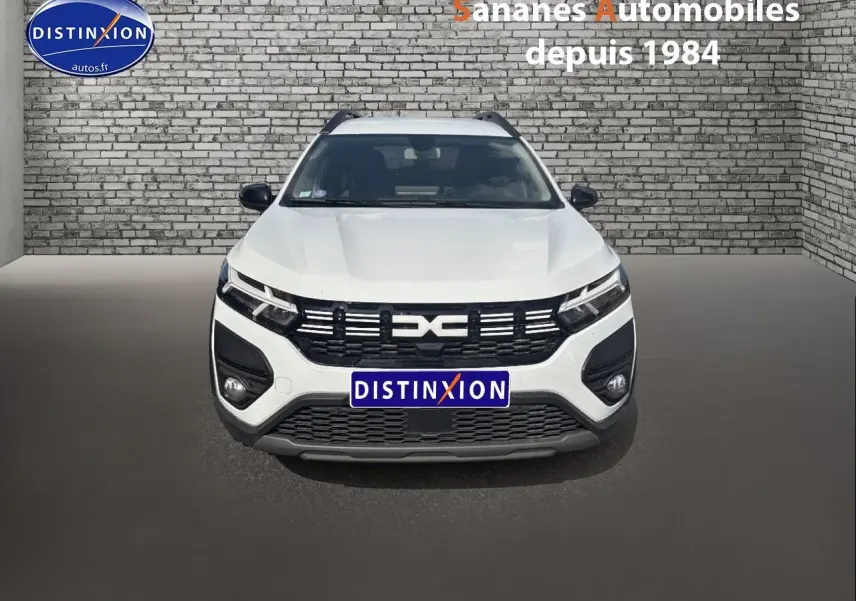 Vue frontale d'un Dacia Jogger blanc avec calandre noire et logo distinctif, stationné devant un mur en briques grises.