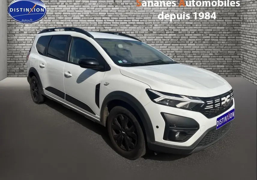 Dacia Jogger blanc vue 3/4 avant gauche avec jantes noires et barres de toit sur fond de mur en briques.