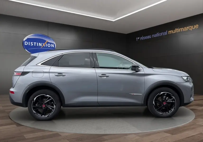 Vue de profil côté droit d'un DS7 Crossback gris clair avec jantes noires et détails chromés sur fond intérieur showroom.
