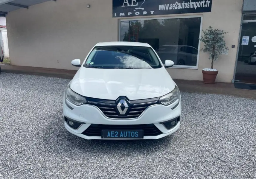 Vue frontale d'une Renault Mégane IV blanche, avec calandre chromée et phares LED, stationnée devant un garage.