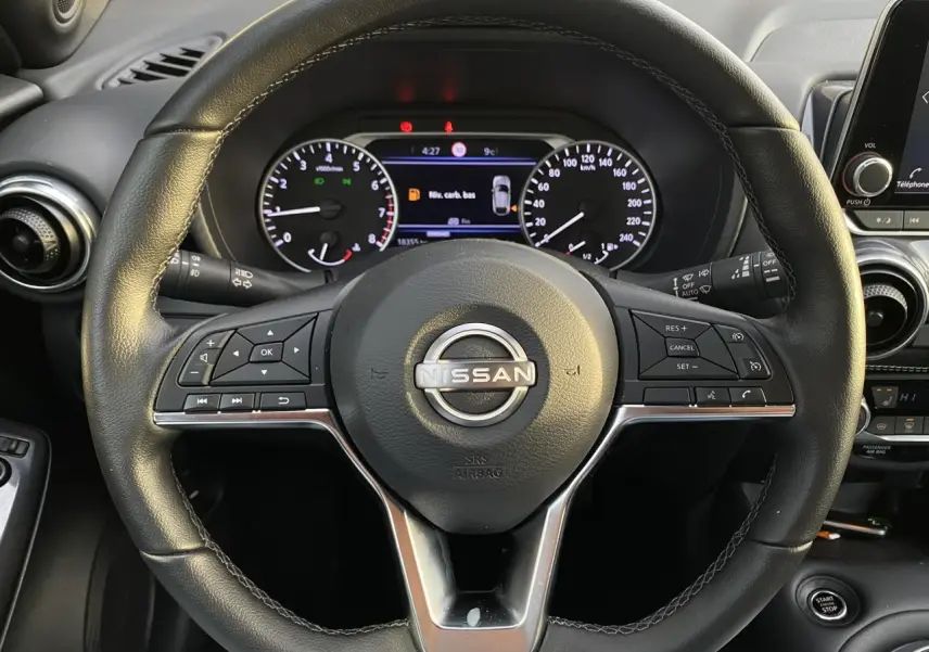 Vue rapprochée du volant noir du Nissan Juke 2024 avec commandes intégrées et tableau de bord numérique en arrière-plan.