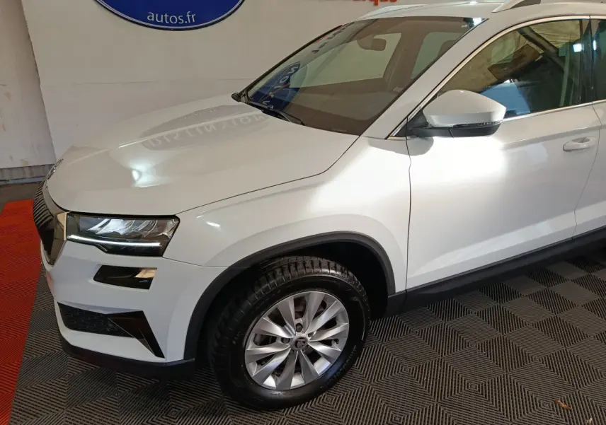 Vue 3/4 avant droit d’un Skoda Karoq blanc 2024 avec jantes alliage et rétroviseurs blancs sur fond intérieur.