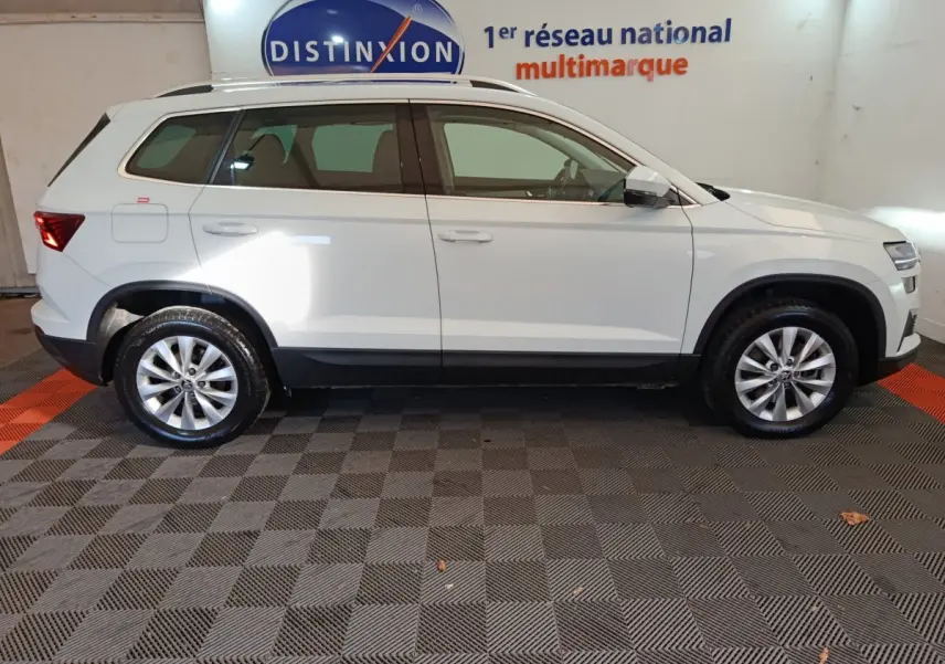 Vue profil côté gauche d'un Skoda Karoq blanc 2024 sur sol damier, avec jantes alliage et rétroviseurs électriques.