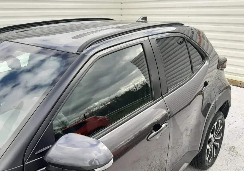 Vue de côté droit avant d'une Toyota Yaris Cross 2024 gris atlas métallisé avec rétroviseur et barres de toit noires.