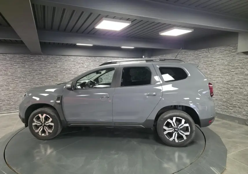 Vue de profil côté gauche d'un Dacia Duster 2024 gris urbain avec jantes alliage 17 pouces et barres de toit.