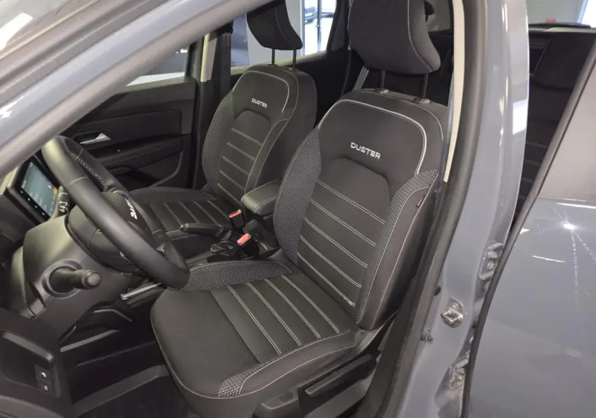 Vue intérieure côté conducteur du Dacia Duster gris 2024, sièges noirs avec surpiqûres blanches et logo Duster brodé.