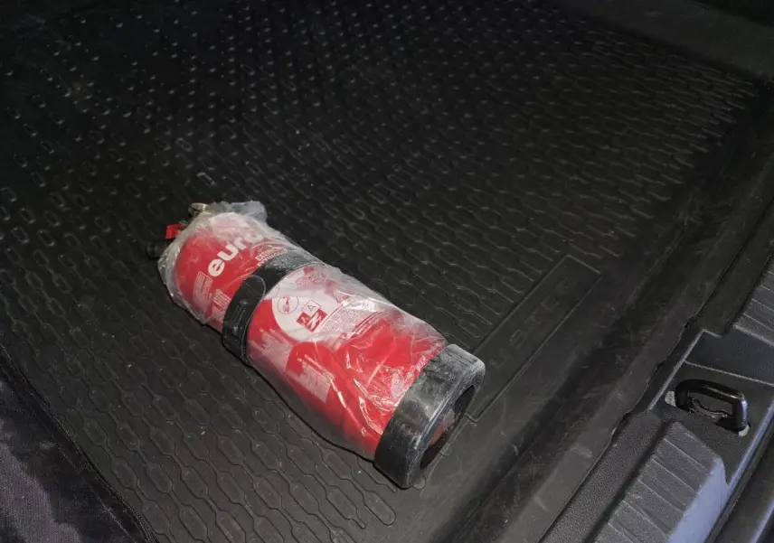 Extérieur coffre noir avec tapis caoutchouc marqué Duster et extincteur rouge posé dessus.