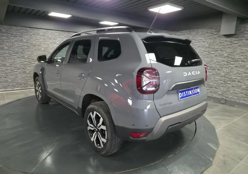Vue 3/4 arrière droit du Dacia Duster 2024 gris urbain avec jantes alliage et barres de toit gris mégalithe.