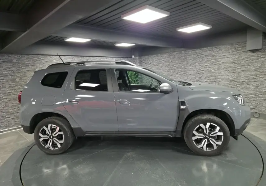 Vue de profil droit du Dacia Duster 2024 gris urbain avec jantes alliage 17 pouces et barres de toit gris.