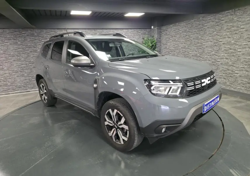 Vue 3/4 avant droit d'un Dacia Duster 2024 gris urbain avec jantes alliage et barres de toit gris mégalithe.
