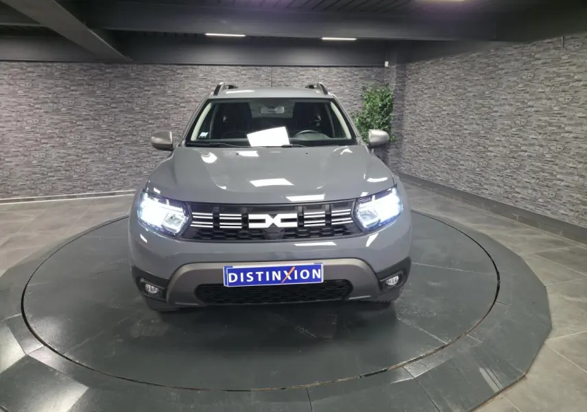 Vue avant d'un Dacia Duster 2024 gris urbain avec calandre noire et phares allumés en intérieur.