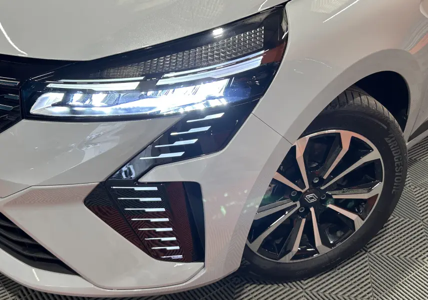 Gros plan sur l'avant droit gris Rafale de la Renault Clio V E-Tech avec phares LED allumés et jante noire brillante.