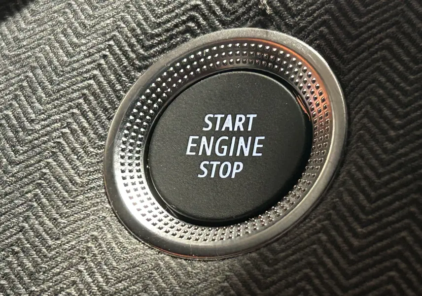 Gros plan sur le bouton Start Engine Stop entouré d’un cerclage métallique, sur un revêtement tissu gris foncé à motif chevrons.