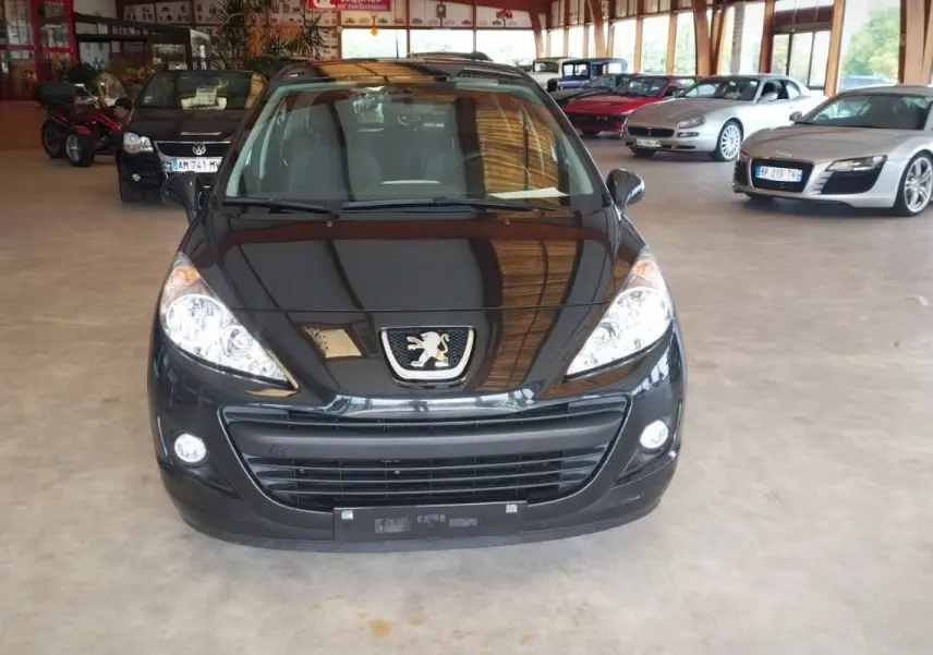 Vue avant d'une Peugeot 207+ noire de 2013, avec phares allumés, stationnée dans un showroom lumineux.