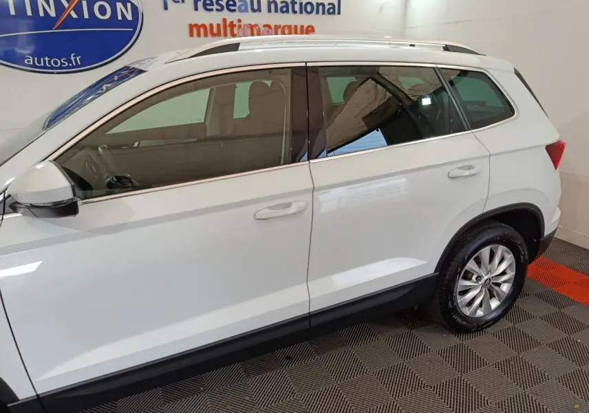 Vue latérale droite d'un Skoda Karoq blanc 2024 avec jantes alliage et barres de toit noires visibles en intérieur showroom.